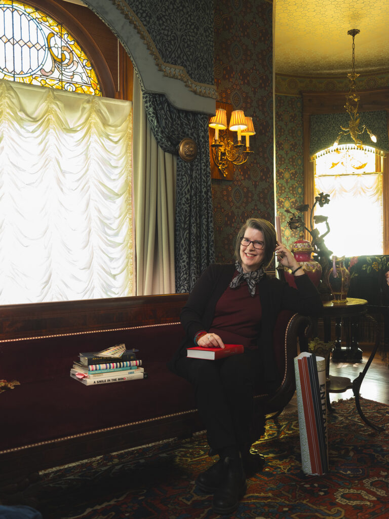 Anne McLaughlin sitting on a Victorian era velvet tufted sofa in front of a window with elegant sheer and heavy drapes
