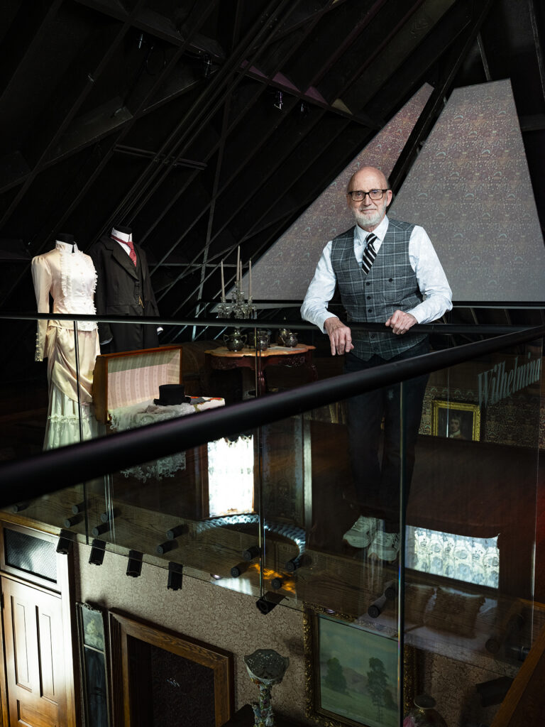 Brad Belk standing with hands on the railing of a glass barrier flanked by mannequins in Victorian garb, reflections of a Victorian decorated bedroom showing in the glass